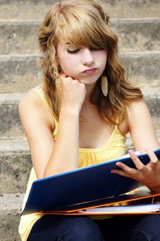 Unhappy or frustrated young blond teenager girl going back to school or looking at her homework in a binder.
