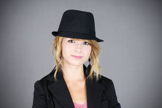 Cute smiling young blond teenager girl model wearing a black felt hat and coat, studio shot over grey background.