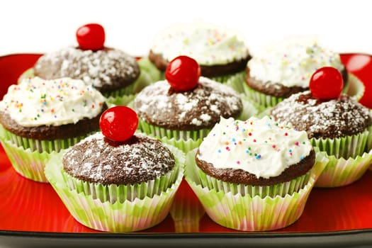 Delicious chocolate cupcakes topped with powdered sugar and maraschimo cherry or with frosting and candies in red plate.
