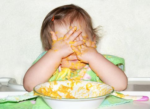 Funny baby with food covered face by putting her hands over her eyes to play picaboo during mealtime, wrong time to do so.