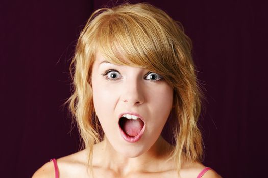 Very surprised or shocked young blond teenager girl making funny face, with eyes and mouth wide open, studio shot.