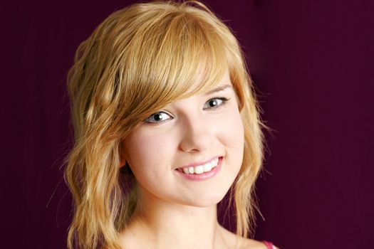 Portrait of cute and friendly yound blong teenager girl smiling, studio shot over deep red or purple background.