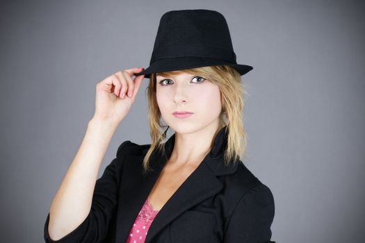 Cute young blond teenager girl model wearing a black felt hat and coat, studio shot over grey background.