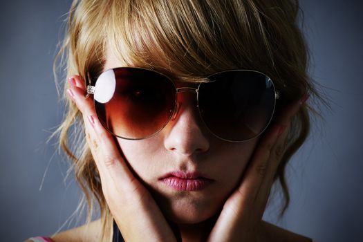 Portrait of a pretty young blond teenager girl with large drak sunglasses, looking sombre, holding her face or covering her ears, over dark grey background