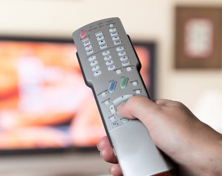 Silver modern TV remote control being pressed by thumb with out of focus screen background