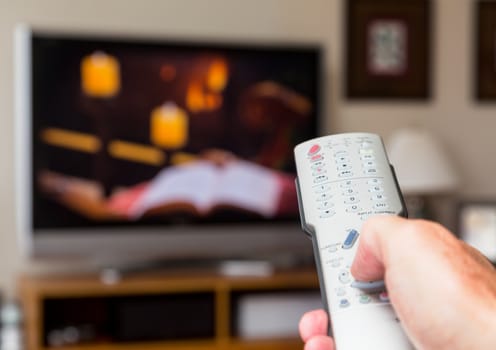 Silver modern TV remote control being pressed by thumb with out of focus screen background