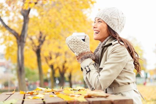Fall concept - autumn woman drinking coffee on park bench under fall foliage. Beautiful young modern woman smiling happy and cheerful in trench coat.