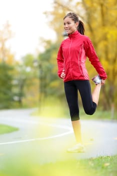 Runner woman stretching thigh after running in city forest park in fall. Beautiful young mixed race Asian / Caucasian female fitness model in autumn color.