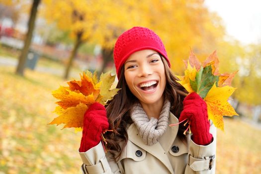 Fall woman happy and bliss in autumn city forest park holding colorful fall leaves smiling happy and joyful wearing tuque and knit gloves. Pretty multiracial Asian and Caucasian girl model.