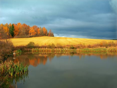 Fall landscape with field after harvest and a pond in the foreground at sunset and dramatic sky