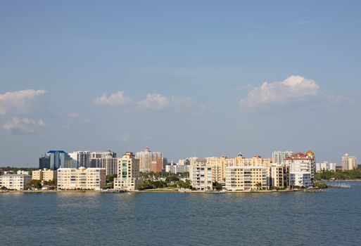 View of buildings on the edge of  Sarasota Bay, Sarasota, Florida from above the water with palm trees, blue sky and white clouds