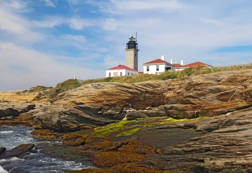 The Beavertail Light lighthouse near Jamestown on Conanicut Island, Rhode Island, viewed from the rocky coast with a bright blue sky and white clouds