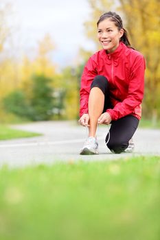 Runner woman tying running shoes outside in fall. Beautiful young fitness model smiling happy in casual jogging clothing.