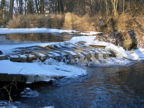            River and ruins of bridge are covered by icicles and lit by a sun