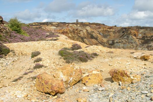 Copper ore rich in colour on the ground in front of an opencast excavation with building in the distance against a blue cloudy sky.