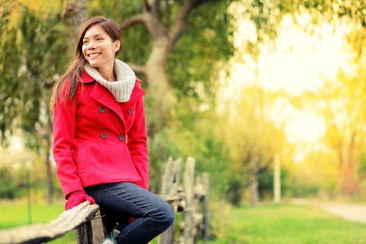 Happy fall woman sitting on countryside fence smiling joyful in autumn forest. Young beautiful multicultural Caucasian / Asian woman model outdoors