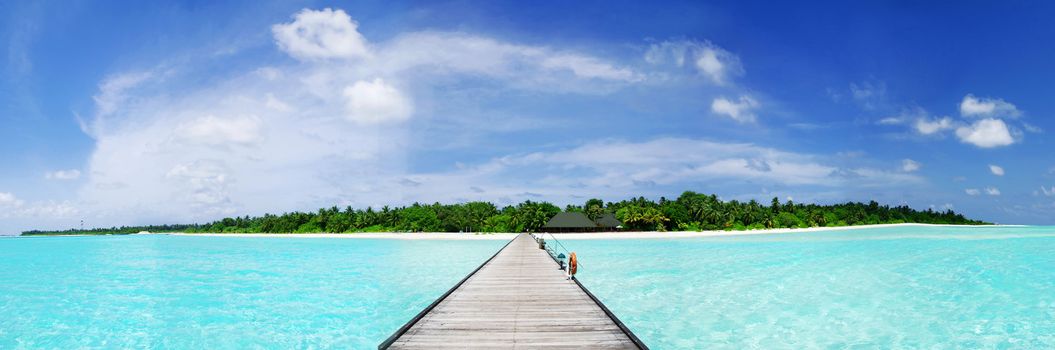 Tropical Maldivian paradise - a jetty leading to a beautiful tropical atoll hiden in the azure Indian ocean. Wide panoramic photograph. 