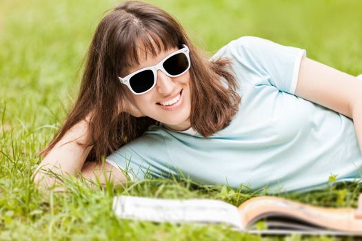 Beauty young smiling caucasian woman in sunglasses reading book outdoor on green grass field