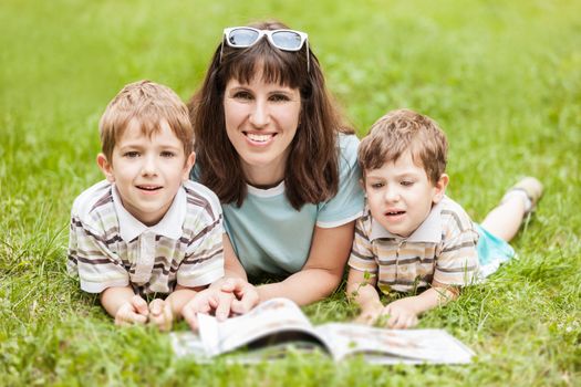 Beauty smiling mother and little sons reading book outdoor on green grass field