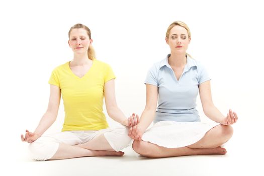 Two attractive blonde women sitting side by side in the lotus position on the floor meditating together isolated on white 