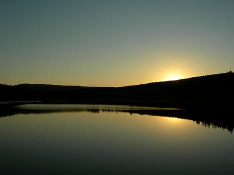           Sun is setting behind forest an is mirroring in the lake