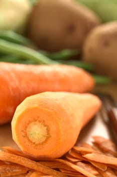 Raw peeled carrot with peeler and other vegetables (green beans and potatoes) on wooden board (Selective Focus, Focus on the middle front part of the carrot)