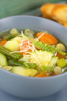 Bowl full of fresh homemade vegetarian Italian minestrone soup made of green beans, zucchini, carrots, potatoes, leek and shell pasta with grated cheese on top and toasted bread in the back (Selective Focus, Focus in the middle of the soup)