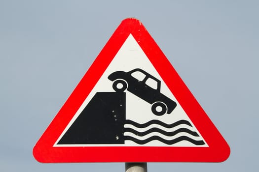 A triangular traffic warning sign with a red border and the symbol for a car driving off a dock wall into the water.