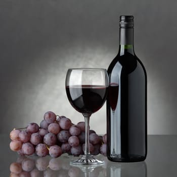 A full bottle of red wine, a glass of wine and black grapes in a wicker basket on a grey background