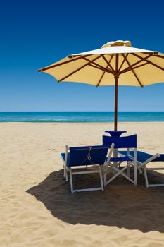 Deck chairs under an umbrella in the sand against the blue sea