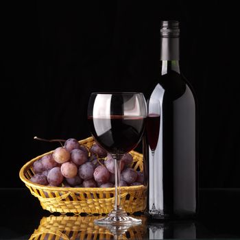 A full bottle of red wine, a glass of wine and black grapes in a wicker basket on a black background