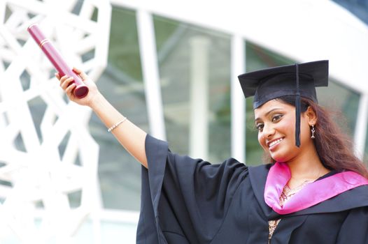 Happy young Indian in a graduation gown and cap holding her diploma