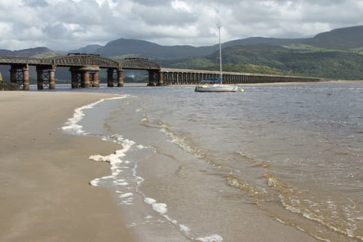 A railway bridge spans an estuary with the sea up on the sand and a is moored in the water backed by dark mountains.