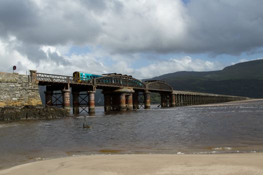 A railway bridge spans an estuary with the sea up on the sand and a train emerges on metal supports backed by dark mountains.