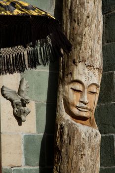A face carved in a log with the casting of a demon on an adjacent wall under an umbrella.