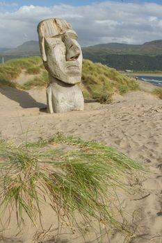 The face of a man carved into a section of wood placed onto a sand dune with grasses and mountains in the background.