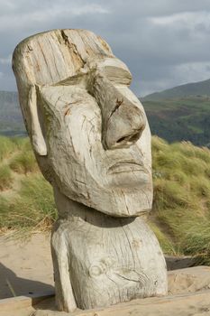 The face of a man carved into a section of wood placed onto a sand dune with grasses and mountains in the background.