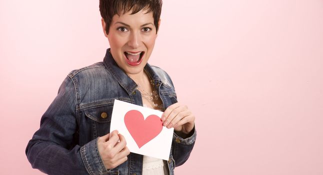 A pretty woman against pink hold her valentine's card with a heart on it