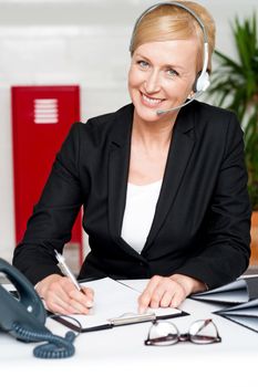 Smiling woman wearing headset writing on notepad sitting in office