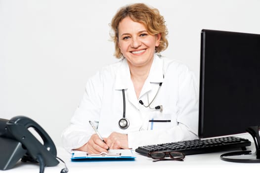 Smiling middle aged female doctor sitting in clinic writing on notepad