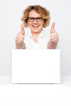 Joyful senior executive showing double thumbs up to camera with laptop kept on desk