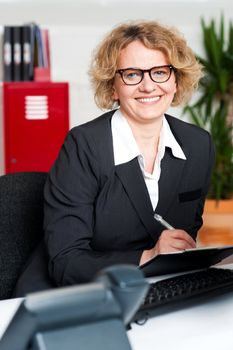 Smiling female executive writing notes on clipboard looking at you