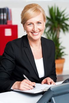 Seated female secretary writing on notepad in office and looking at you