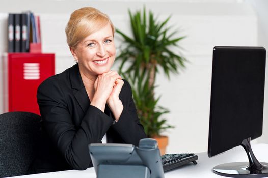 Smiling aged businesswoman sitting with hands on chin looking at camera