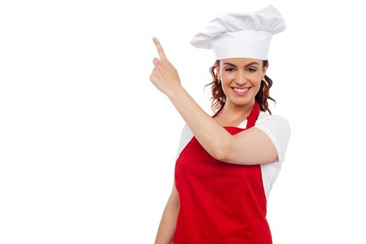 Attractive young beautiful woman chef wearing red apron and toque pointing at copy space