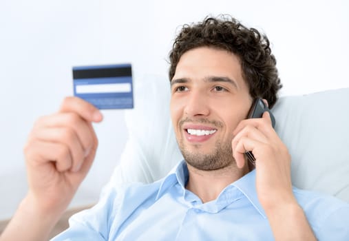 Young handsome man looking on credit card and talking on modern mobile phone. Studio shot.