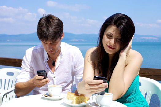 Happy couple texting with mobile phone during breakfast with cappuccino and cake on the lake