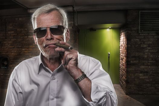 Retired man with strong personality standing in the darkness in sunglasses puffing on a big cigar, quintessential stereotype of a retired successful executive