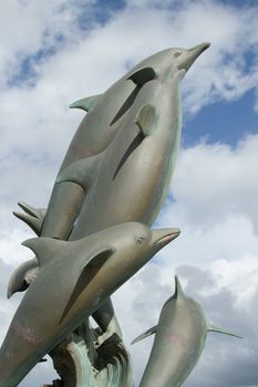 A metal cast, moulded, statue of a pod, group of dolphins against a blue sky with clouds.