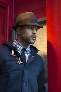 Young black man with fedora standing in red doorway looking outward toward the street.